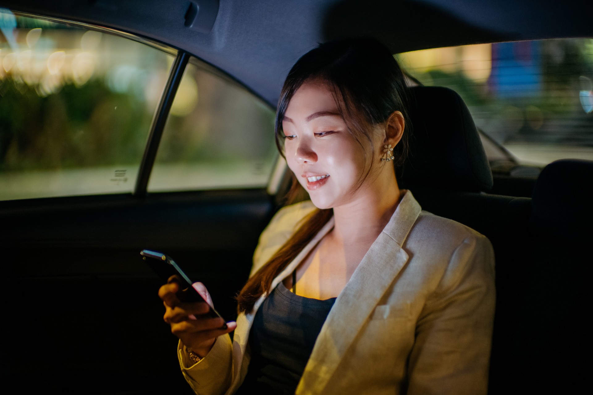 Femme d’affaires chinoise asiatique utilisant le téléphone intelligent s’asseyant au siège d’auto de passager dans une ville de nuit